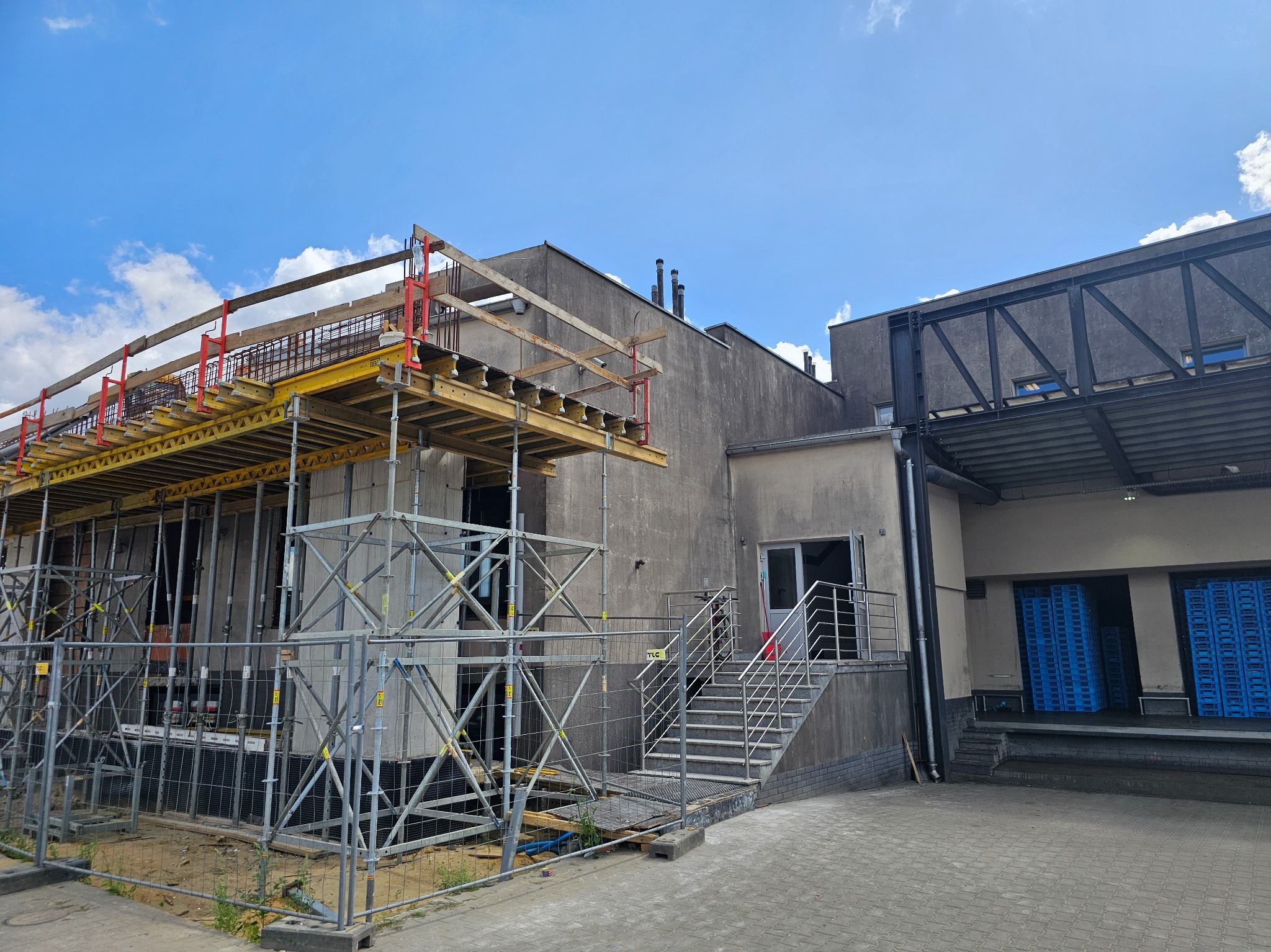 Budynek w trakcie rozbudowy z widocznymi rusztowaniami i elementami konstrukcyjnymi. Metalowe schody prowadzą do wejścia. Błękitne palety w tle.