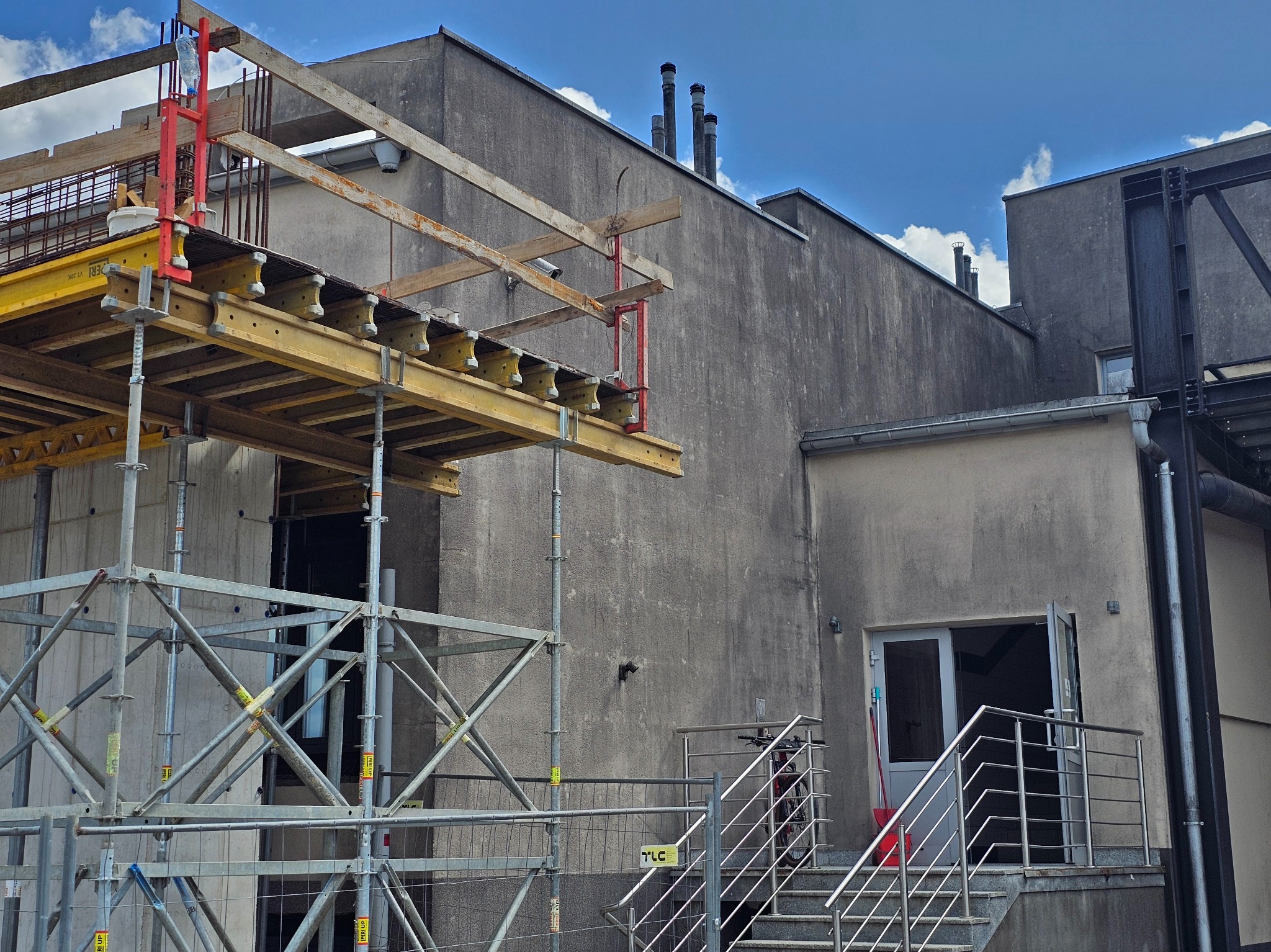 Szare budynki w trakcie budowy z widocznymi rusztowaniami i elementami konstrukcyjnymi. Na schodach rower. Błękitne niebo z chmurami w tle.