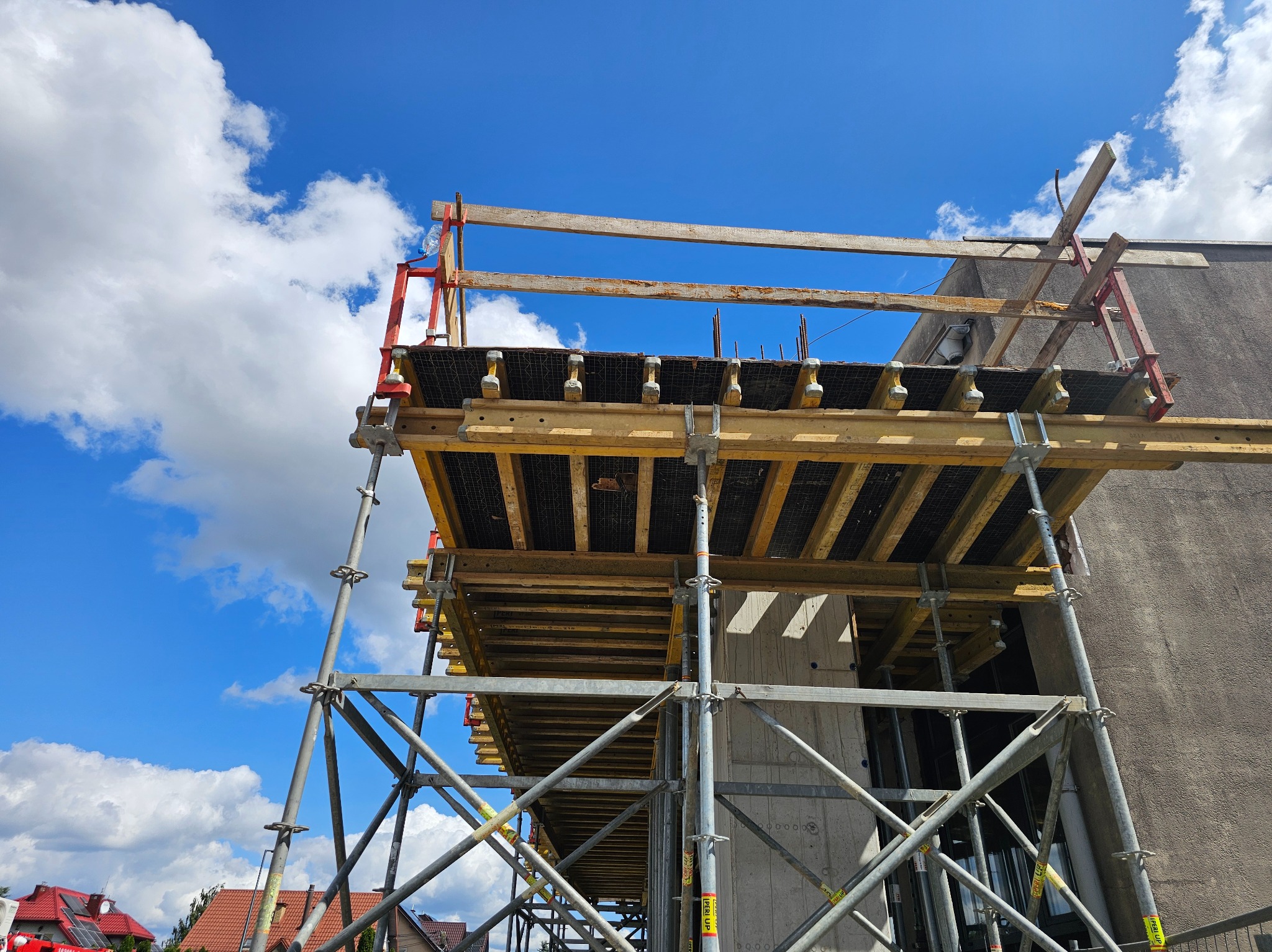 Szare rusztowanie z żółtymi elementami szalunkowymi na tle błękitnego nieba z chmurami. Widoczny fragment betonowej konstrukcji budynku w trakcie budowy.