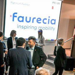 Film&Foto Beata Pietruszka - Uczestnicy konferencji w kuluarach rozmawiają, na ekranie wyświetlony logotyp firmy Faurecia, w tle widoczny baner z hasłem 'Become a modern engineer'.