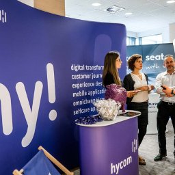 Film&Foto Beata Pietruszka - Stoisko firmy Hycom na targach: cztery osoby rozmawiają na tle ścianki z logo i hasłami, na stoliku fioletowa owca i miska z cukierkami.