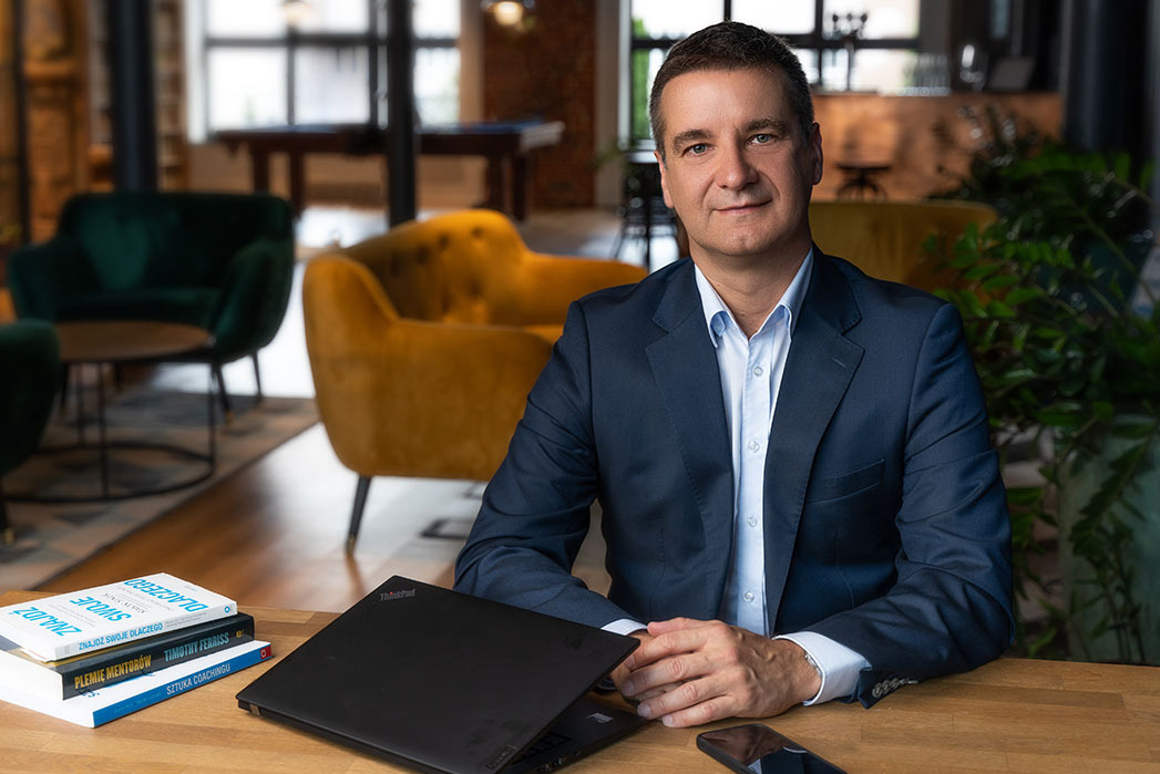 Portret biznesowy mężczyzny w garniturze siedzącego za biurkiem z laptopem i książkami, w tle widoczne fotele i stół bilardowy.