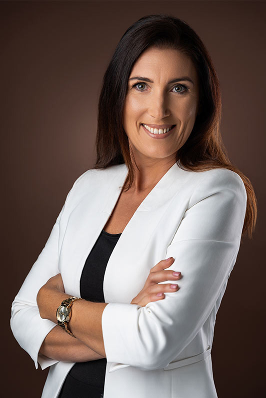 Portret kobiety w białym żakiecie na brązowym tle, z założonymi rękami i złotym zegarkiem, uśmiechająca się do kamery.