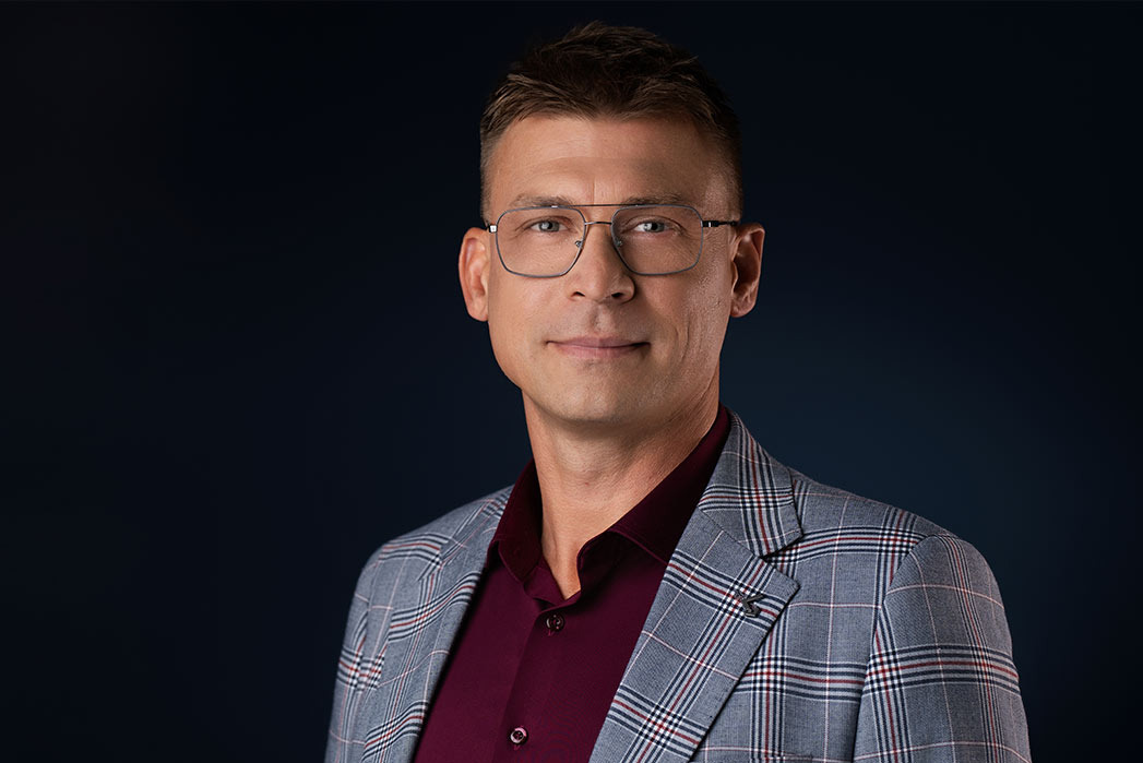 Portret mężczyzny w okularach i marynarce w kratę na ciemnym tle, z subtelnym uśmiechem.