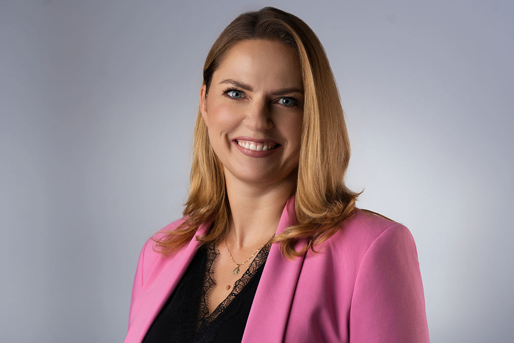 Portret uśmiechniętej kobiety w różowej marynarce i czarnej bluzce z koronką, na jednolitym, jasnoszarym tle.