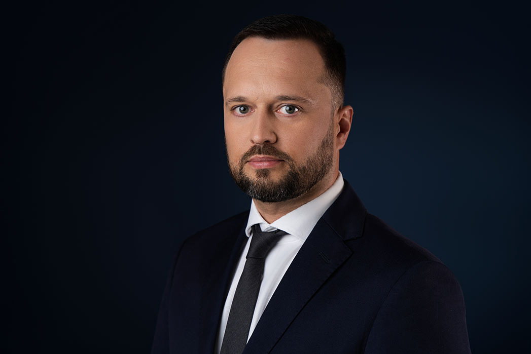 Portret biznesowy mężczyzny w ciemnym garniturze i białej koszuli z czarnym krawatem na ciemnoniebieskim tle.