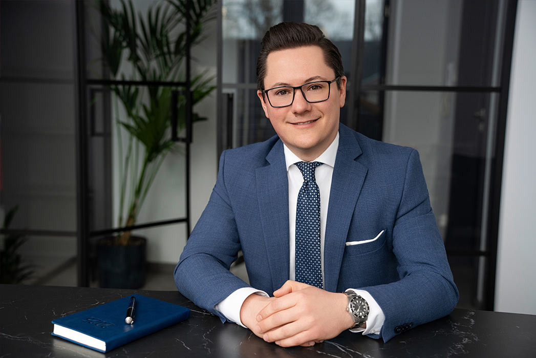 Portret biznesowy uśmiechniętego mężczyzny w niebieskim garniturze, siedzącego za czarnym biurkiem z kalendarzem i piórem, w tle roślina w doniczce i przeszklona ściana.