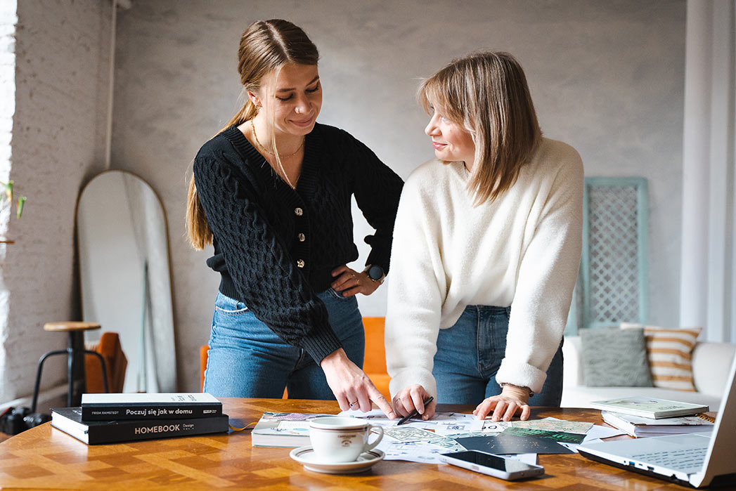 Dwie kobiety w swetrach pochylają się nad stołem z rozłożonymi planami, kubkiem kawy i laptopem, analizując projekt wnętrza w jasnym pomieszczeniu z lustrem i książkami.