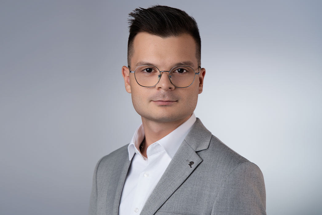 Portret biznesowy mężczyzny w okularach i szarej marynarce z przypinką w kształcie grzyba na klapie, na jednolitym szarym tle.