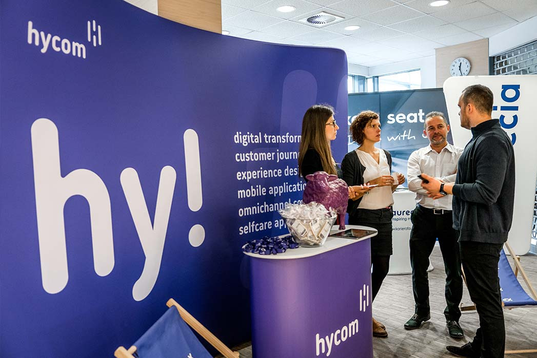 Stoisko firmy Hycom na targach: cztery osoby rozmawiają na tle ścianki z logo i hasłami, na stoliku fioletowa owca i miska z cukierkami.