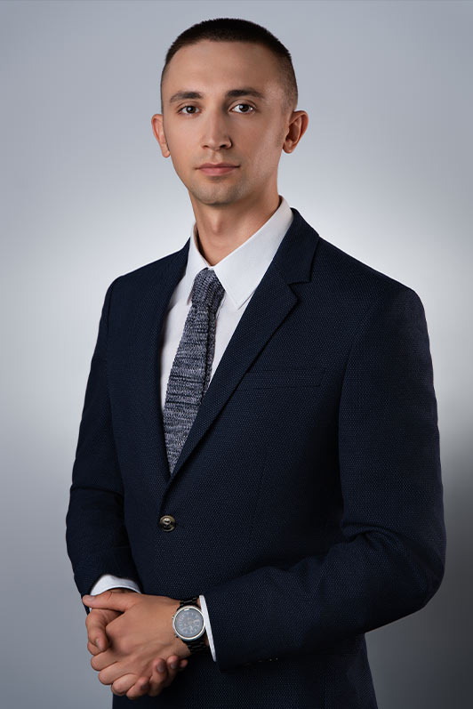 Portret mężczyzny w granatowym garniturze z szarą dzierganą fakturą krawata i zegarkiem na nadgarstku, na jednolitym szarym tle.