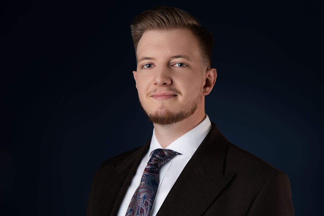 Portret młodego mężczyzny w brązowym garniturze i wzorzystym krawacie na ciemnoniebieskim tle, z delikatnym uśmiechem i starannie ułożoną fryzurą.