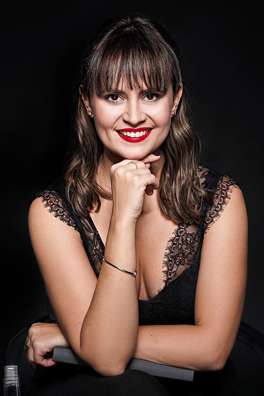 Portret uśmiechniętej kobiety w czarnej koronkowej sukience, z ręką podpartą pod brodą, na ciemnym tle.