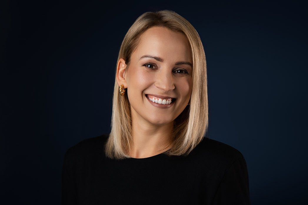 Portret uśmiechniętej kobiety z blond włosami, w czarnej bluzce i złotych kolczykach na ciemnoniebieskim tle.