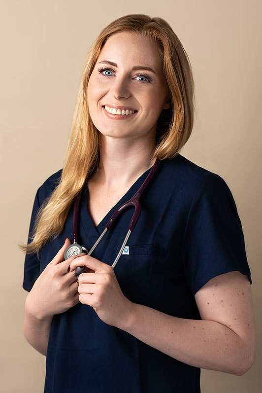 Portret uśmiechniętej blondynki w granatowym uniformie medycznym ze stetoskopem na szyi, na jednolitym beżowym tle.