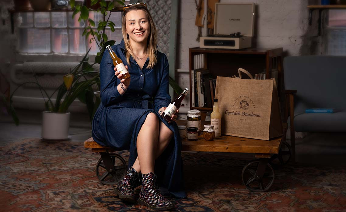 Uśmiechnięta kobieta w dżinsowej sukience siedzi na wózku z kołami, prezentując dwie butelki z napojami marki 'Ogródek Dziadunia', obok torba z logo tej samej firmy, w tle gramofon i książki.