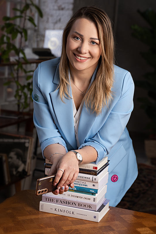 Portret uśmiechniętej kobiety w błękitnej marynarce, opierającej się o stos książek na drewnianym stole, trzymającej smartfon w dłoniach.