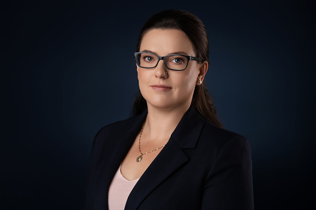 Portret kobiety w okularach, ubrana w ciemną marynarkę i jasną bluzkę, z delikatną złotą biżuterią na ciemnym tle.