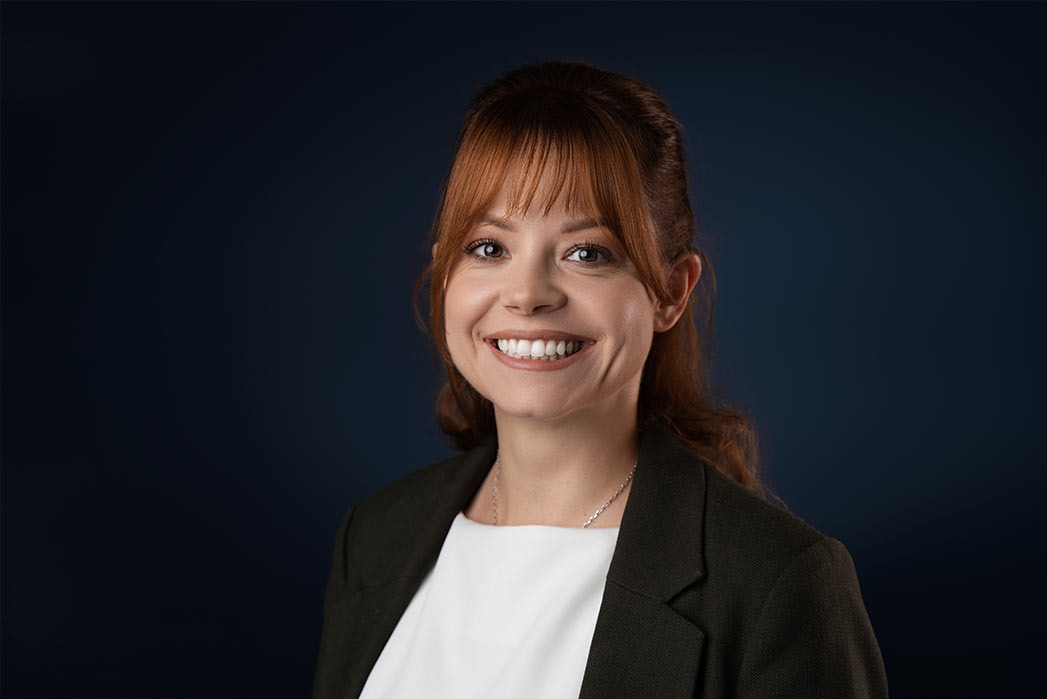 Portret uśmiechniętej kobiety z rudymi włosami, ubrana w białą bluzkę i czarną marynarkę na ciemnoniebieskim tle.