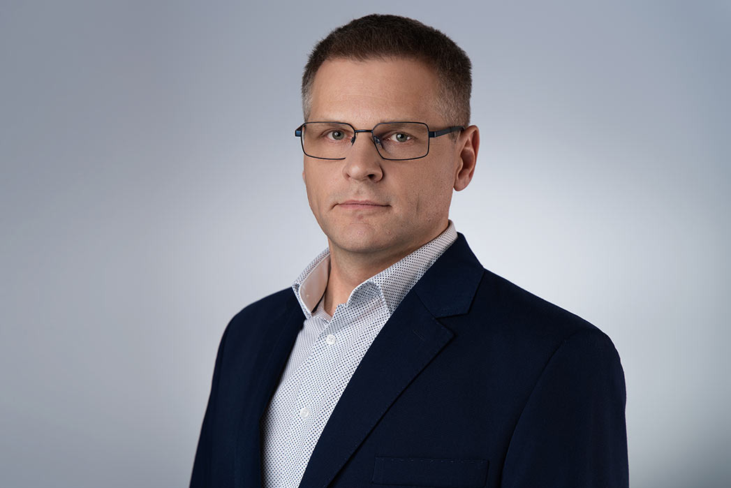 Portret mężczyzny w okularach i granatowej marynarce, na jednolitym, jasnoszarym tle.