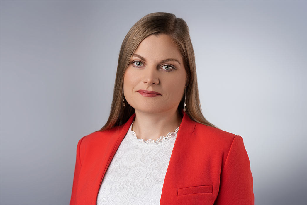 Portret kobiety w czerwonej marynarce i białej bluzce z koronkowym kołnierzykiem, na jednolitym szarym tle.