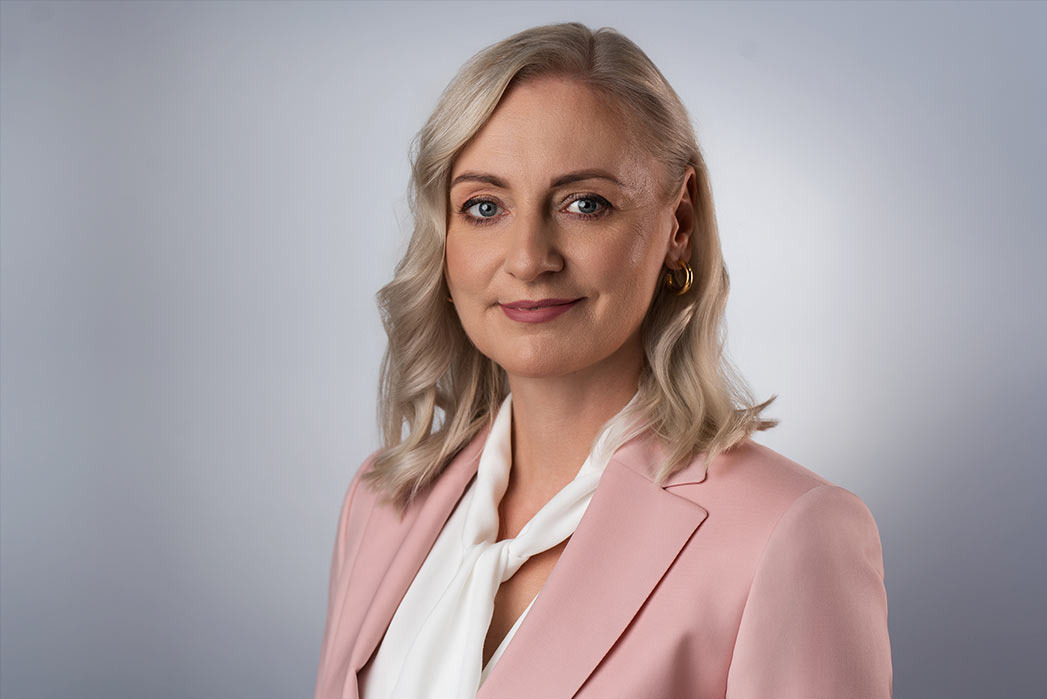 Portret kobiety o blond włosach, ubrana w różowy żakiet i białą bluzkę z wiązaniem, pozująca na szarym tle.