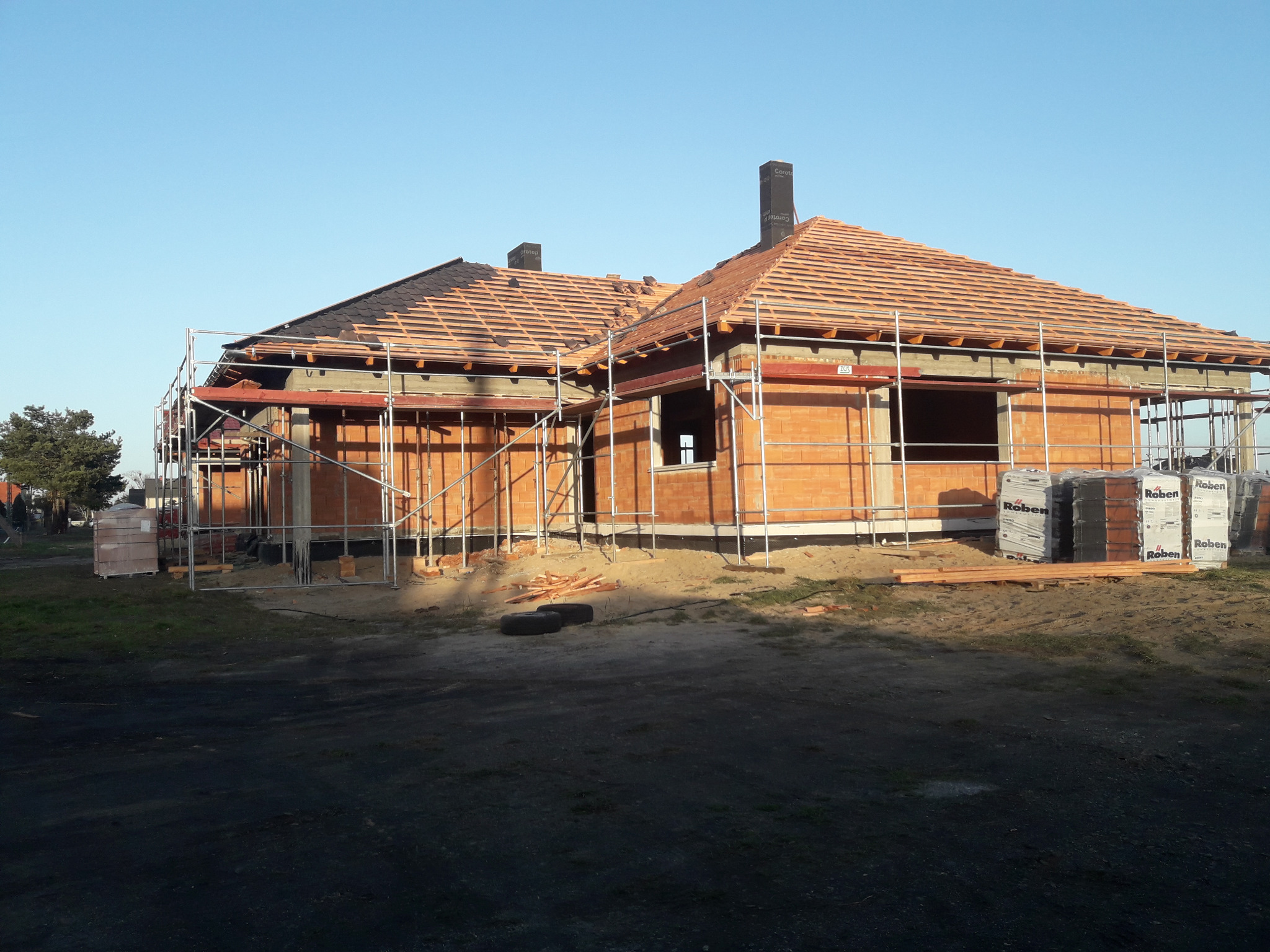 Niedokończony dom z czerwonej cegły z częściowo położonym dachem z dachówki ceramicznej, otoczony rusztowaniami i stertami materiałów budowlanych Roben.