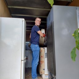 Piotr Jaśkowiak Złota Rączka - Mężczyzna w busie transportuje lodówki, uśmiechnięty, w rękawicach roboczych. Widoczne liście drzewa, materac i wnętrze pojazdu dostawczego.