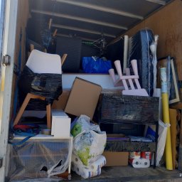 Piotr Jaśkowiak Złota Rączka - Wnętrze załadowanego busa przeprowadzkowego: meble zabezpieczone folią, krzesła, kartony, torby z zawartością, widoczne elementy wyposażenia domu podczas transportu.