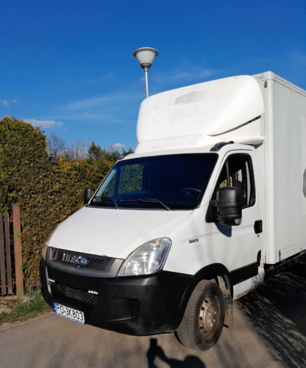 Biały samochód dostawczy Iveco z zabudową kontenerową, zaparkowany na ulicy w słoneczny dzień, widoczny z przodu, z shadow sylwetki fotografa w rogu kadru.