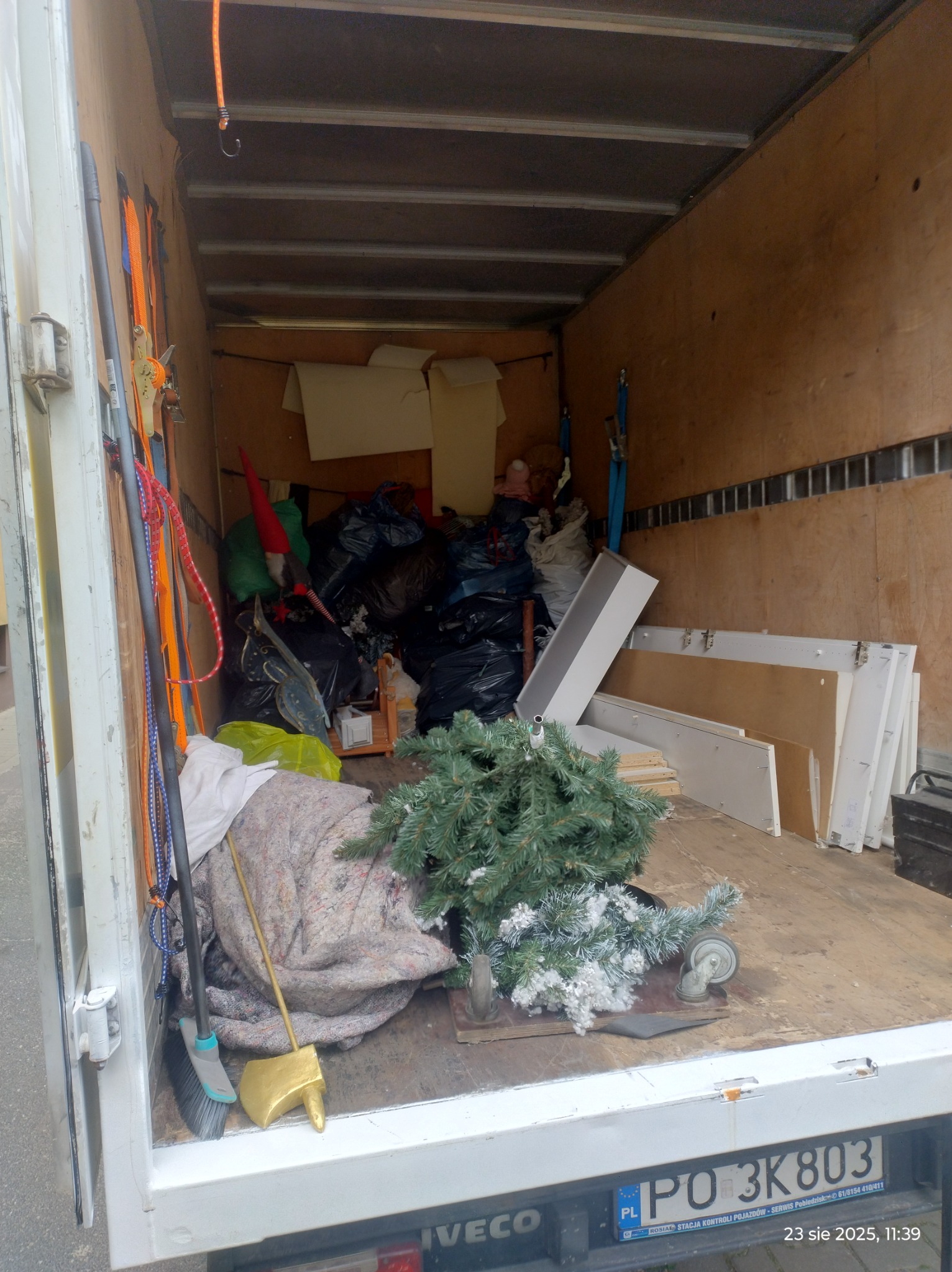 Wnętrze furgonetki załadowane workami, meblami i choinką, gotowe do transportu. Widoczne zabezpieczenia ładunku i tablica rejestracyjna pojazdu.