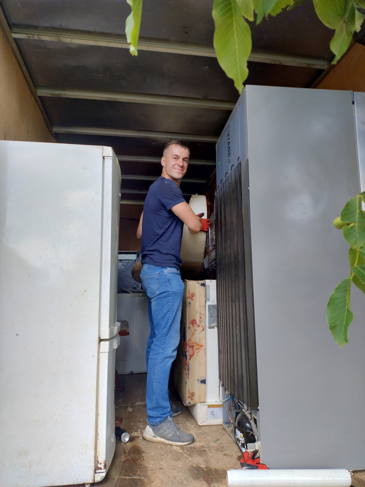 Mężczyzna w busie transportuje lodówki, uśmiechnięty, w rękawicach roboczych. Widoczne liście drzewa, materac i wnętrze pojazdu dostawczego.
