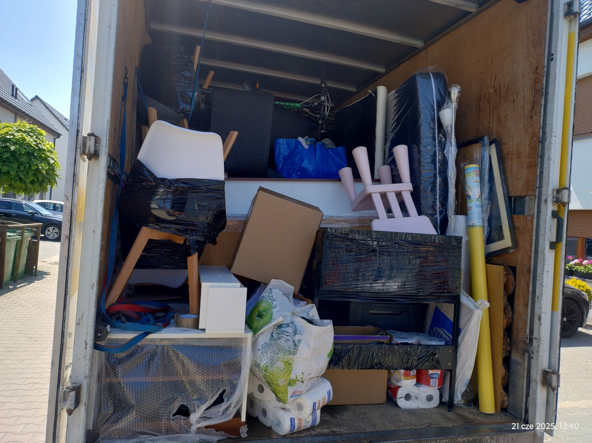 Wnętrze załadowanego busa przeprowadzkowego: meble zabezpieczone folią, krzesła, kartony, torby z zawartością, widoczne elementy wyposażenia domu podczas transportu.