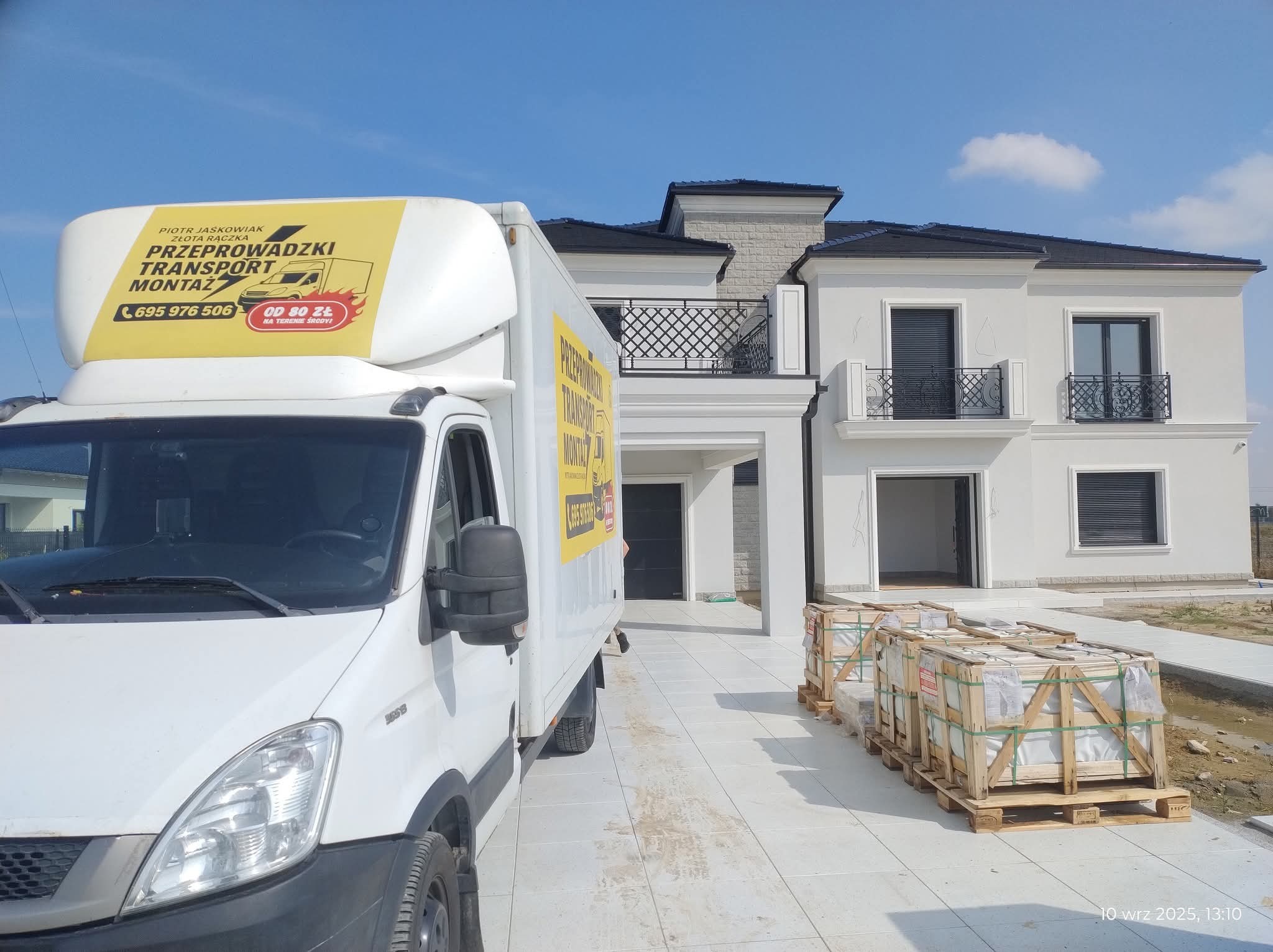 Biała furgonetka z reklamą firmy przeprowadzkowej na tle nowoczesnego domu z balkonami i paletami z materiałami budowlanymi. Słoneczny dzień.