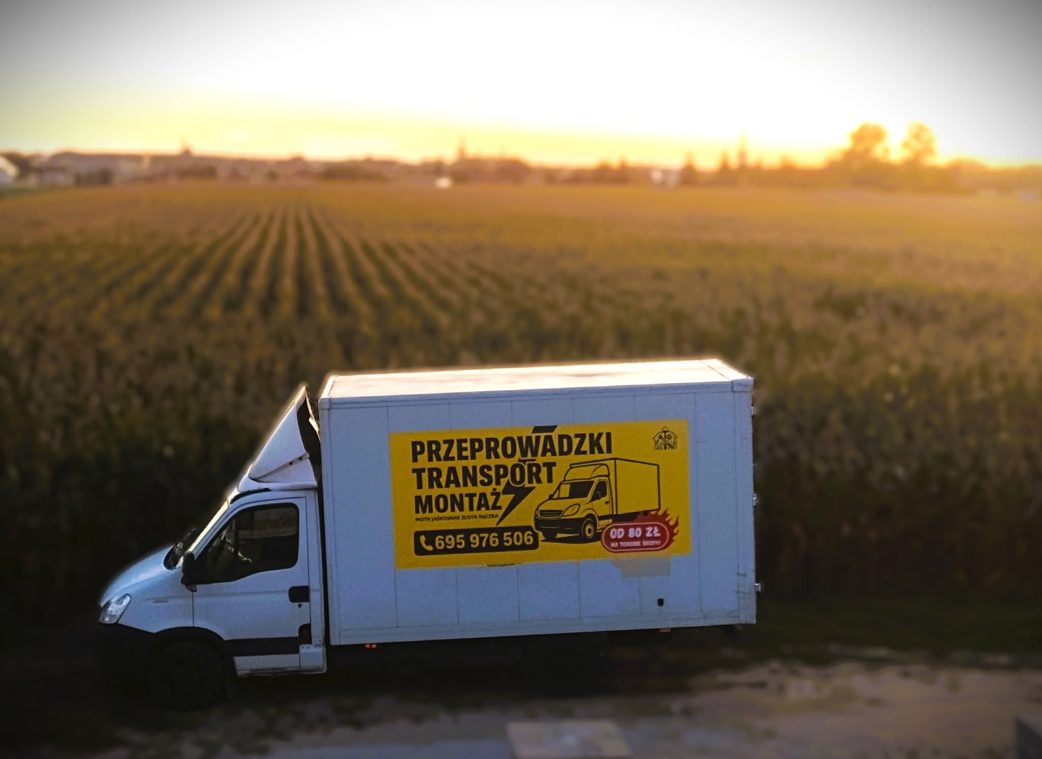 Biały samochód dostawczy z żółtą reklamą usług przeprowadzek, transportu i montażu na tle pola kukurydzy o zachodzie słońca. Widoczny numer telefonu i informacja o cenie.