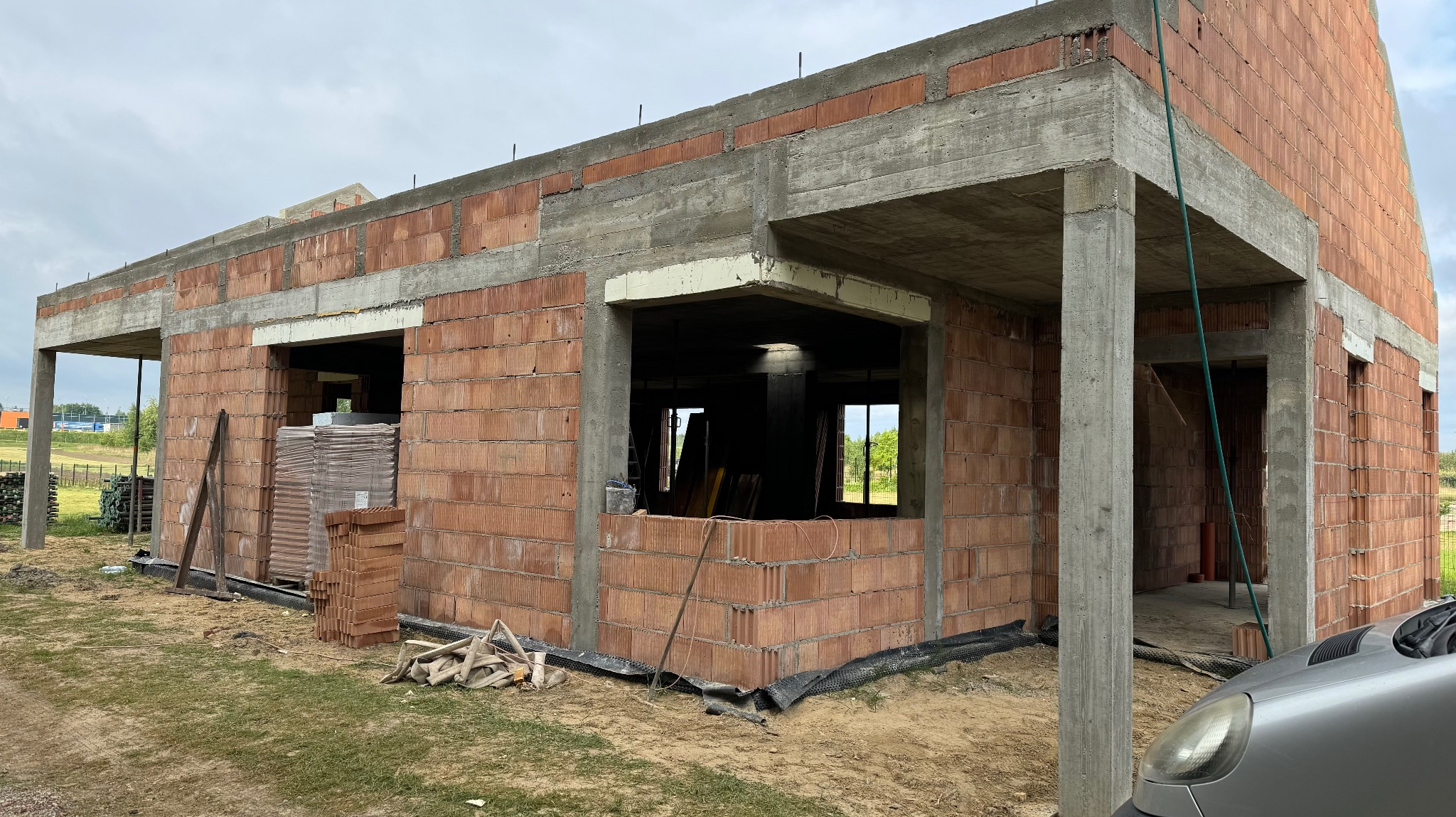 Budowa domu z czerwonej cegły, widoczne betonowe słupy i nadproża. Na pierwszym planie stos cegieł i fragment samochodu. Budowa w trakcie realizacji.