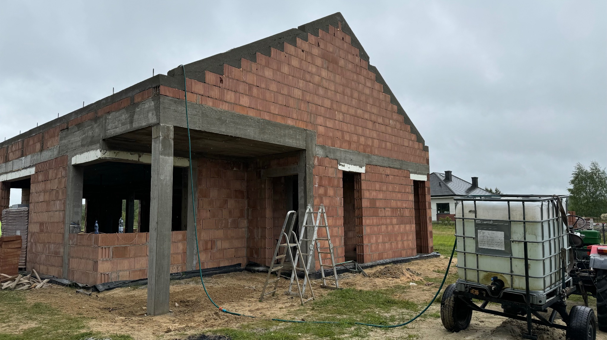 Surowy stan domu z czerwonej cegły, widoczne rusztowania i zbiornik na wodę. Budowa w toku, zarys dachu i otworów okiennych. Tło: pochmurne niebo.
