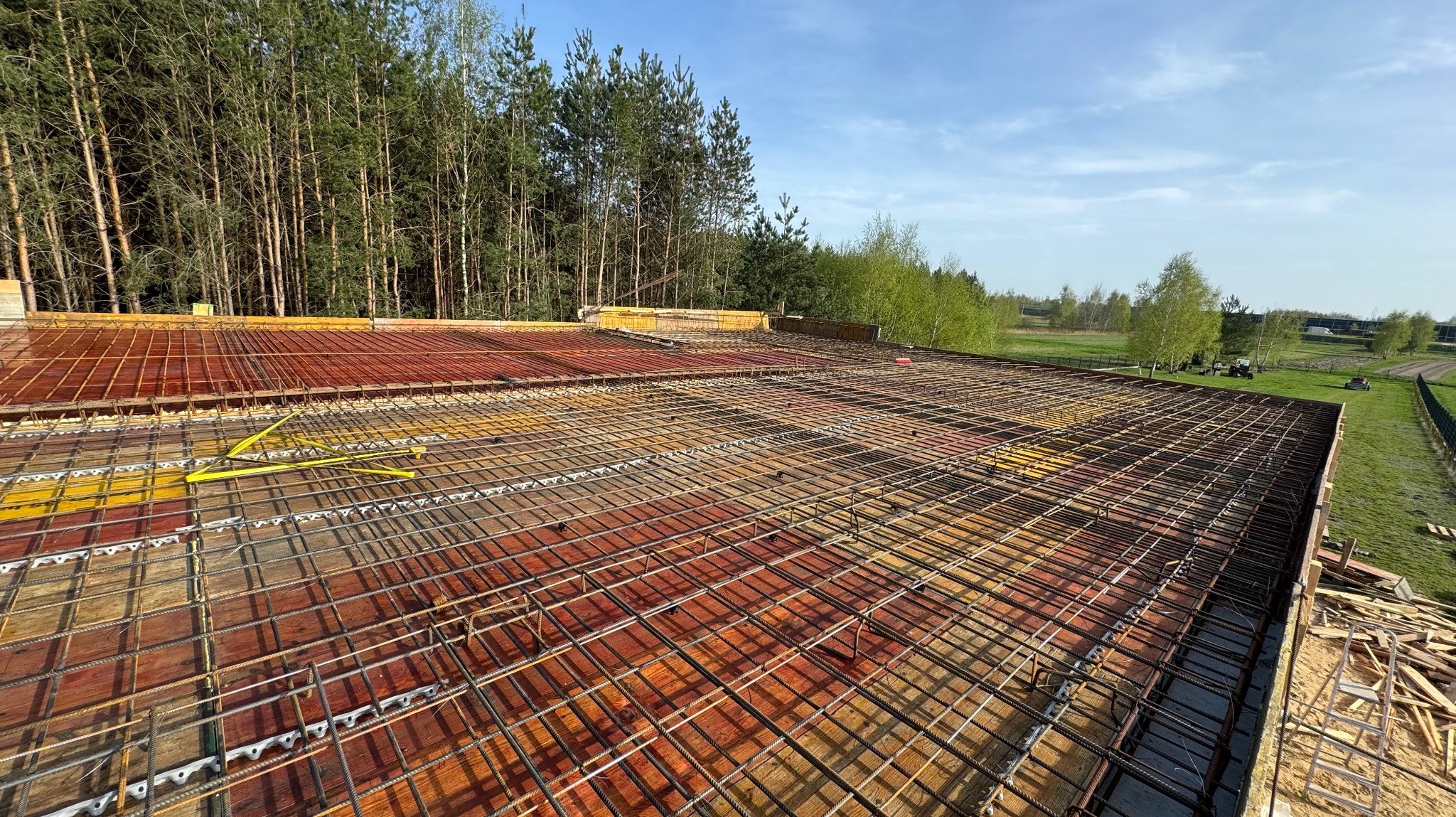 Zbrojenie stropu budynku w trakcie budowy, widok z góry na siatkę z prętów stalowych ułożoną na deskowaniu, w tle las i błękitne niebo.