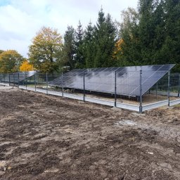 Sebastian Drejka - Panele słoneczne za ogrodzeniem z siatki na betonowej podmurówce, na przygotowanym terenie. Widoczne drzewa w tle i pochmurne niebo.