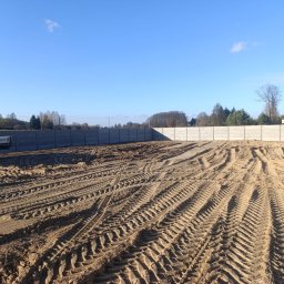 Sebastian Drejka - Przygotowany teren pod budowę z betonowym ogrodzeniem w tle. Widoczne ślady opon i pracownik przy ciężarówce. Błękitne niebo.