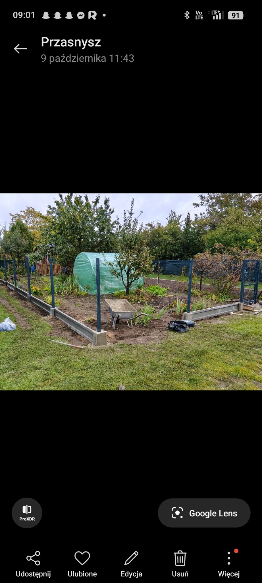 Wykopane fundamenty pod ogrodzenie panelowe z podmurówką prefabrykowaną. Widoczna taczka i szklarnia w tle. Prace ziemne w ogrodzie na etapie budowy ogrodzenia.