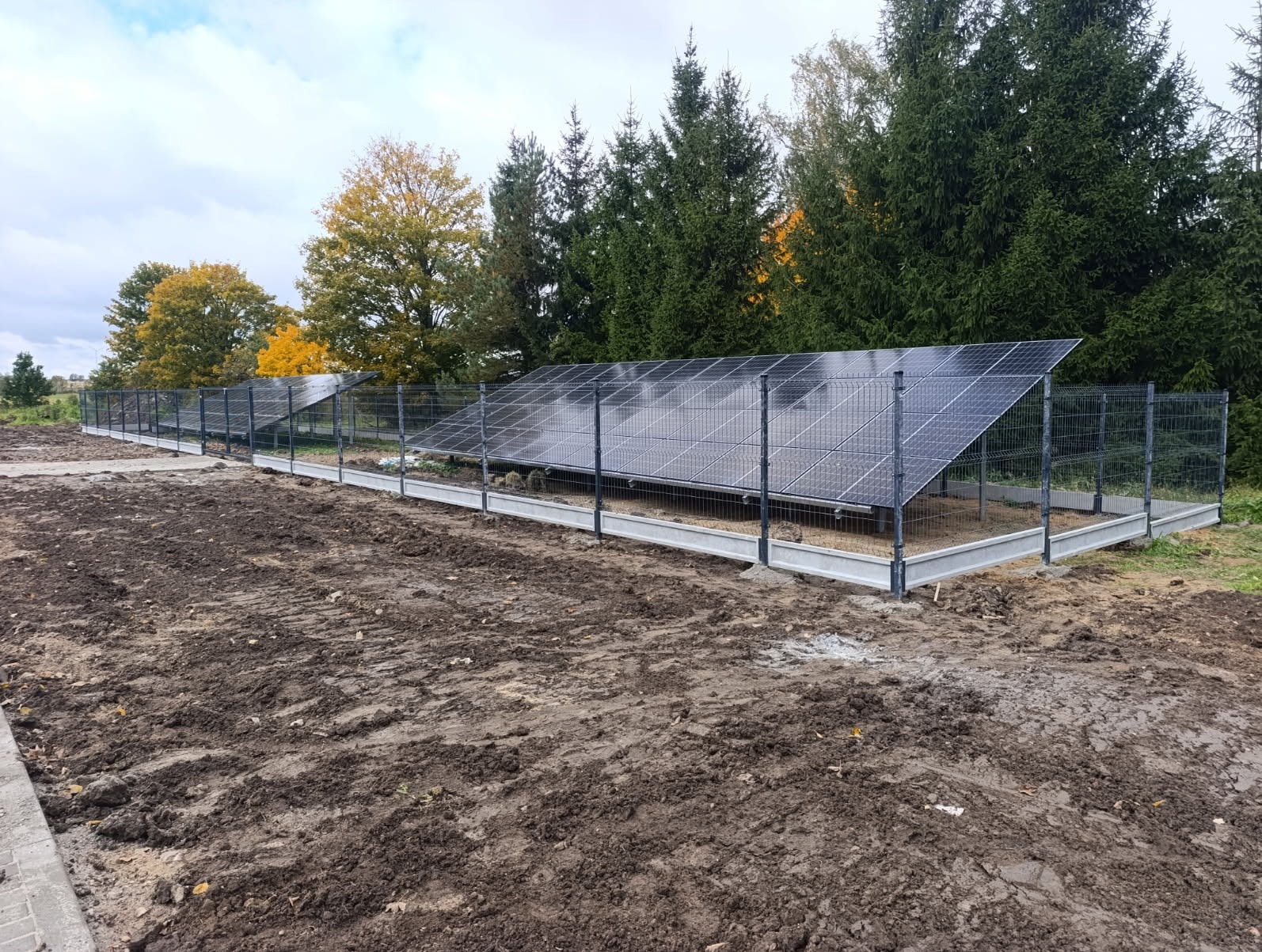 Panele słoneczne za ogrodzeniem z siatki na betonowej podmurówce, na przygotowanym terenie. Widoczne drzewa w tle i pochmurne niebo.