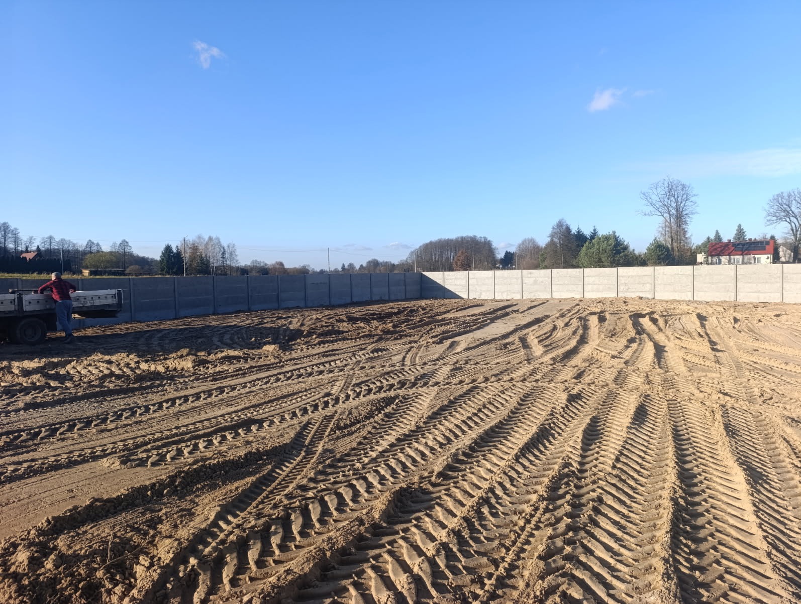 Przygotowany teren pod budowę z betonowym ogrodzeniem w tle. Widoczne ślady opon i pracownik przy ciężarówce. Błękitne niebo.