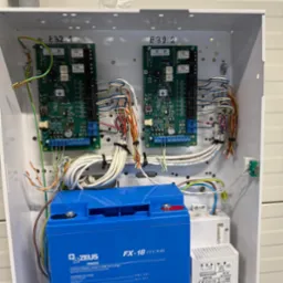 Centrala alarmowa w białej obudowie, widoczne moduły elektroniczne, akumulator Fenix Zeus FX-18 i transformator. Przewody uporządkowane, instalacja wewnątrz skrzynki.