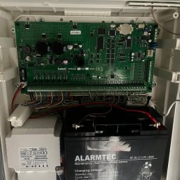 Przemysław Łyko - Centrala alarmowa Satel Integra 128 Plus w obudowie, z widoczną płytą główną, transformatorem Pulsar i akumulatorem Alarmtec, połączona przewodami.
