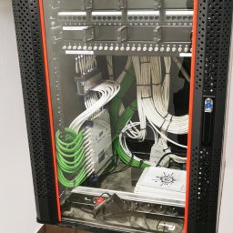 Przemysław Łyko - Szafa rack z okablowaniem strukturalnym: zielone i białe przewody uporządkowane wewnątrz czarnej szafy z pomarańczowymi akcentami, widok z przodu na tle beżowych płytek.