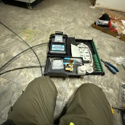 Przemysław Łyko - Otwarta skrzynka z narzędziami i światłowodami na podłodze. Widoczne złącza światłowodowe, spawarka światłowodowa i inne narzędzia. Praca w toku, instalacja telekomunikacyjna.