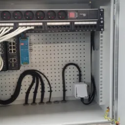 Otwarta szafa elektryczna z listwą zasilającą, patch panelem, switchem i okablowaniem. Widoczne gniazda, kable Ethernet, zasilacz i uziemienie.