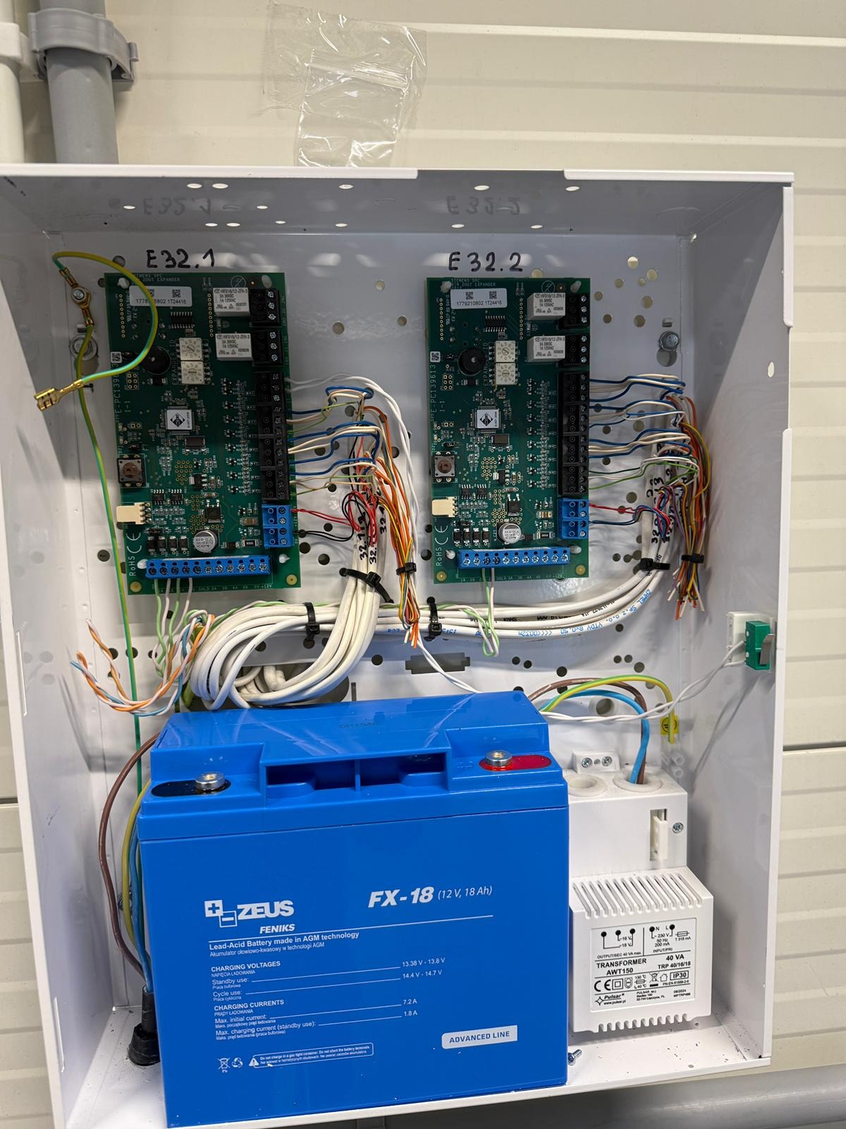 Centrala alarmowa w białej obudowie, widoczne moduły elektroniczne, akumulator Fenix Zeus FX-18 i transformator. Przewody uporządkowane, instalacja wewnątrz skrzynki.