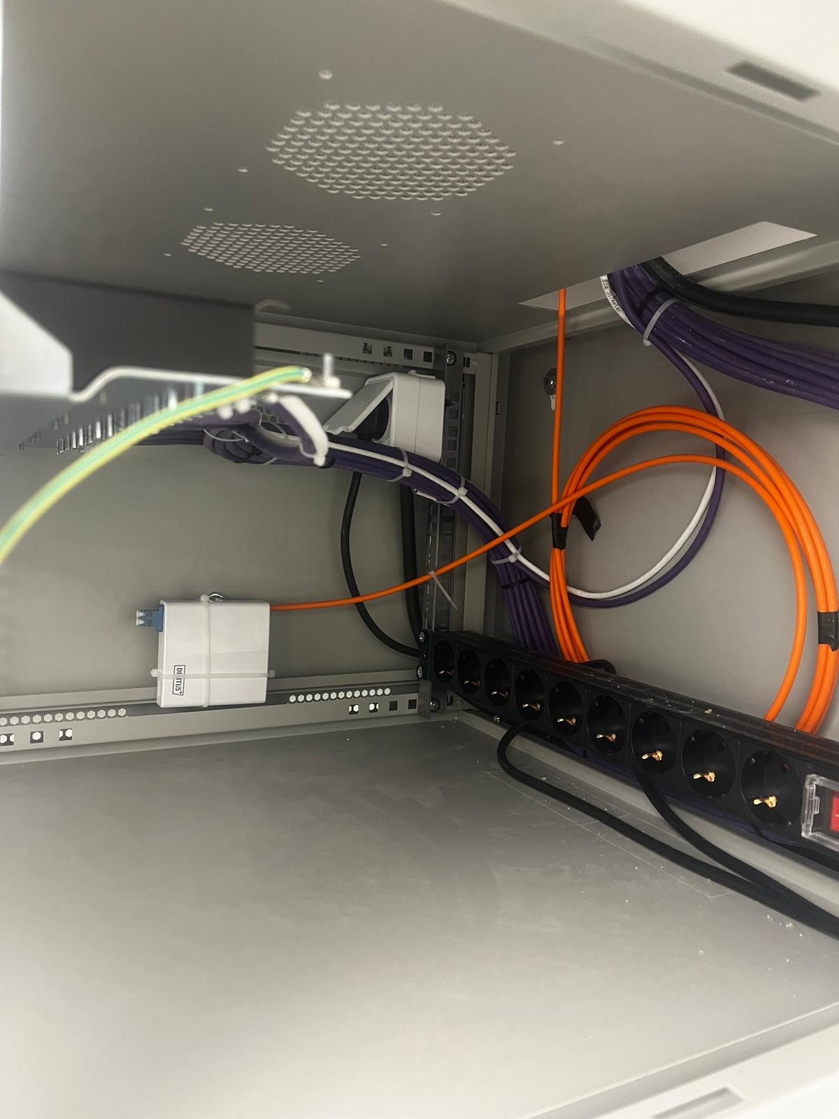 Wnętrze szafy rack z zainstalowaną listwą zasilającą, patch panelem i uporządkowanymi kablami światłowodowymi oraz ethernetowymi w kolorach pomarańczowym i fioletowym.